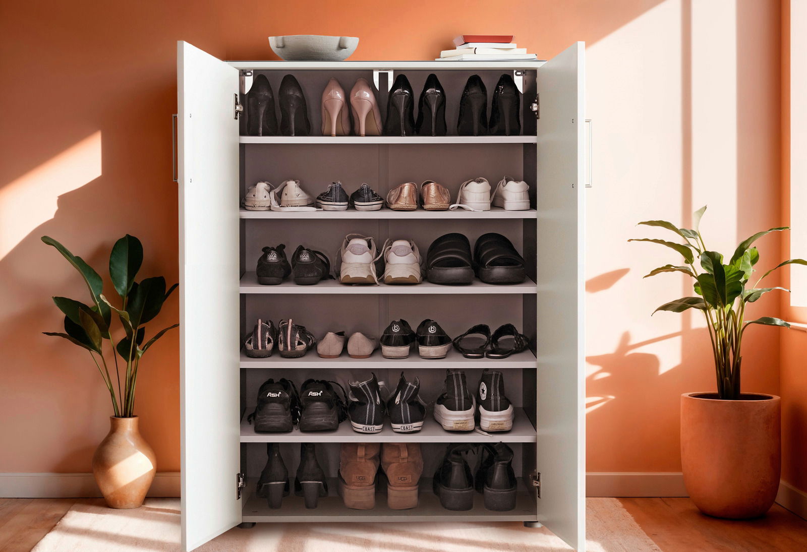 Schuhschrank mit offenen Türen, gefüllt mit verschiedenen Schuhen, in einem hellen Raum mit Pflanzen, aus frontaler Perspektive.