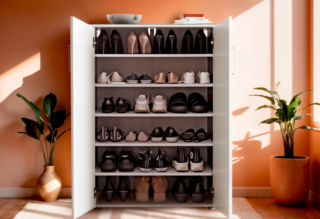 Schuhschrank mit offenen Türen, gefüllt mit verschiedenen Schuhen, in einem hellen Raum mit Pflanzen, aus frontaler Perspektive.