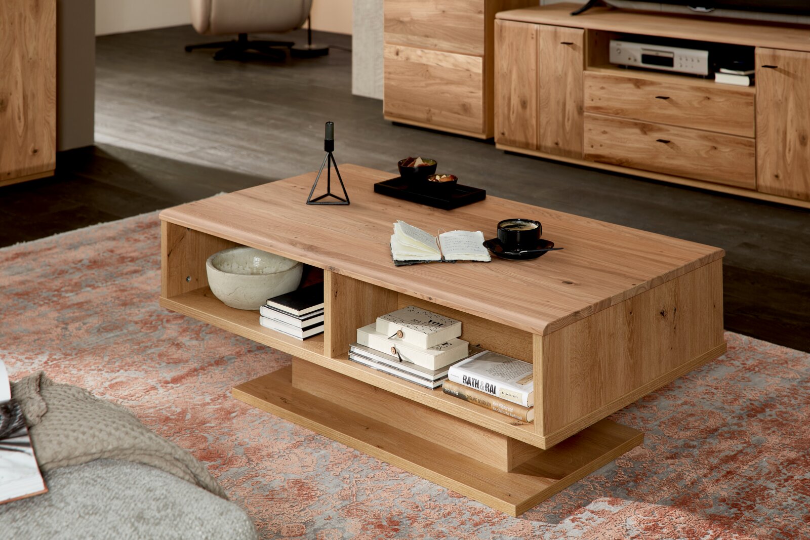 Cantus Couchtisch REIMS Moderner rechteckiger Couchtisch aus hellem Massivholz mit offenem Ablagefach und breiter Bodenplatte, im Wohnzimmer auf gemustertem Teppich dekoriert, Perspektive: schräg von vorne leicht von oben.