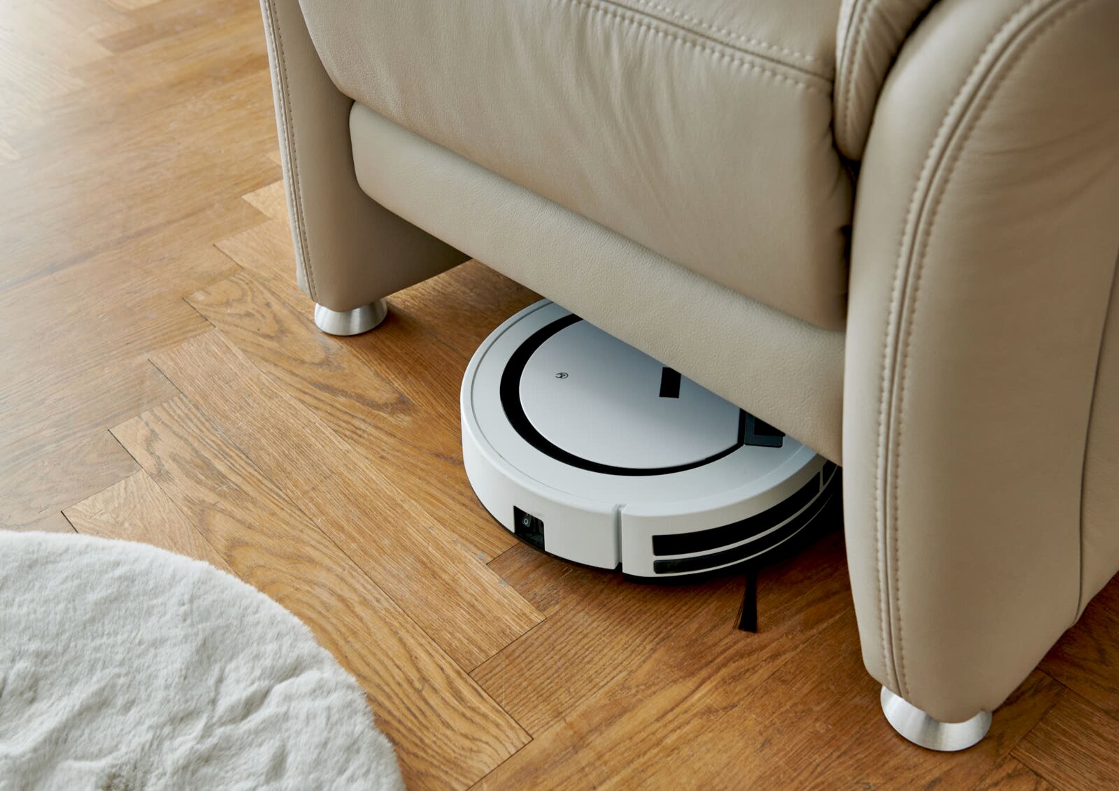 Een robotstofzuiger maakt de houten vloer schoon onder een beige leren fauteuil, gezien vanuit de zijkant.