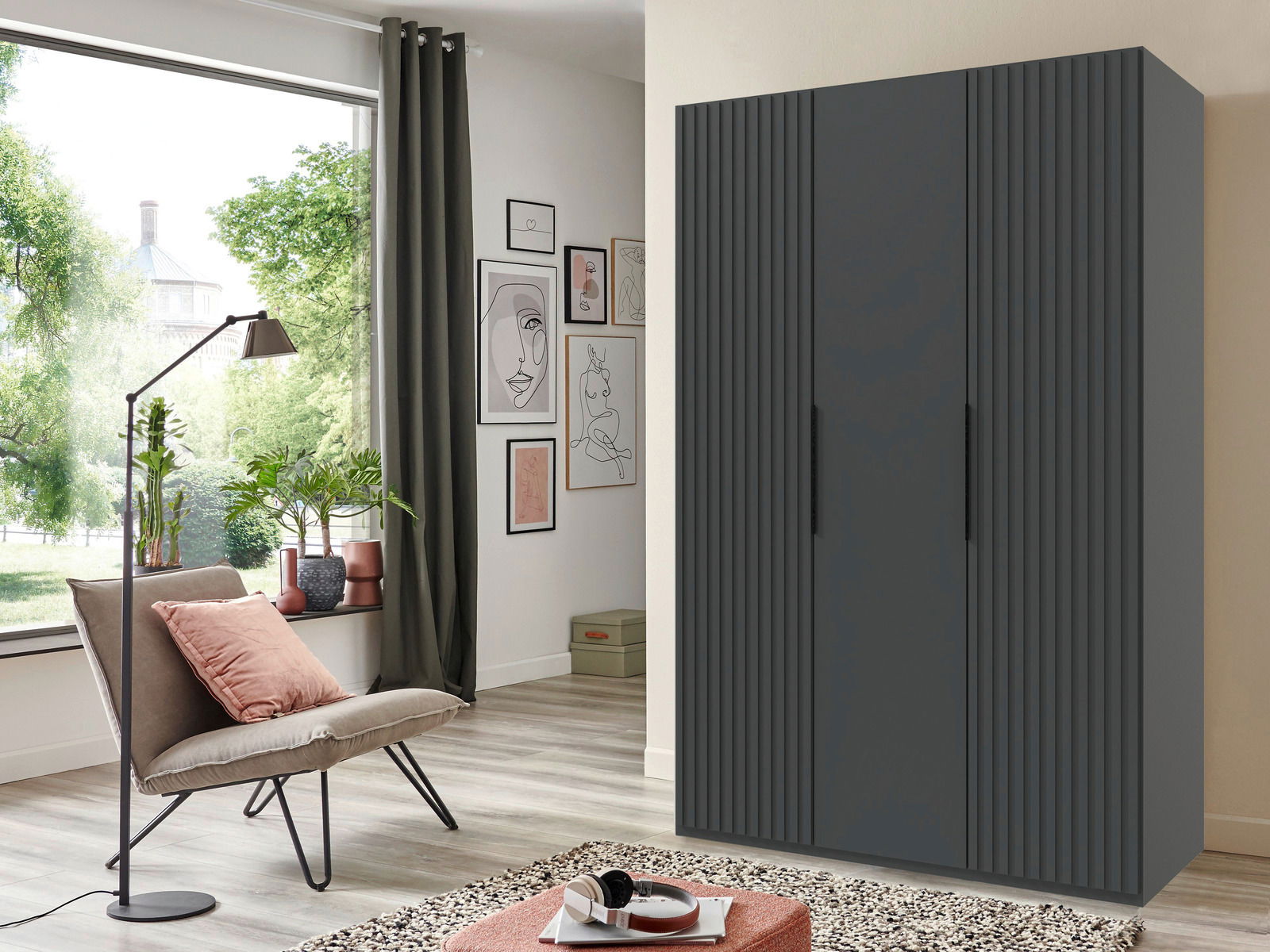 Moderner dreitüriger Drehtürenschrank in dunklem Grau, frontal fotografiert in einem stilvoll eingerichteten Wohnzimmer mit großen Fenstern und grüner Aussicht.