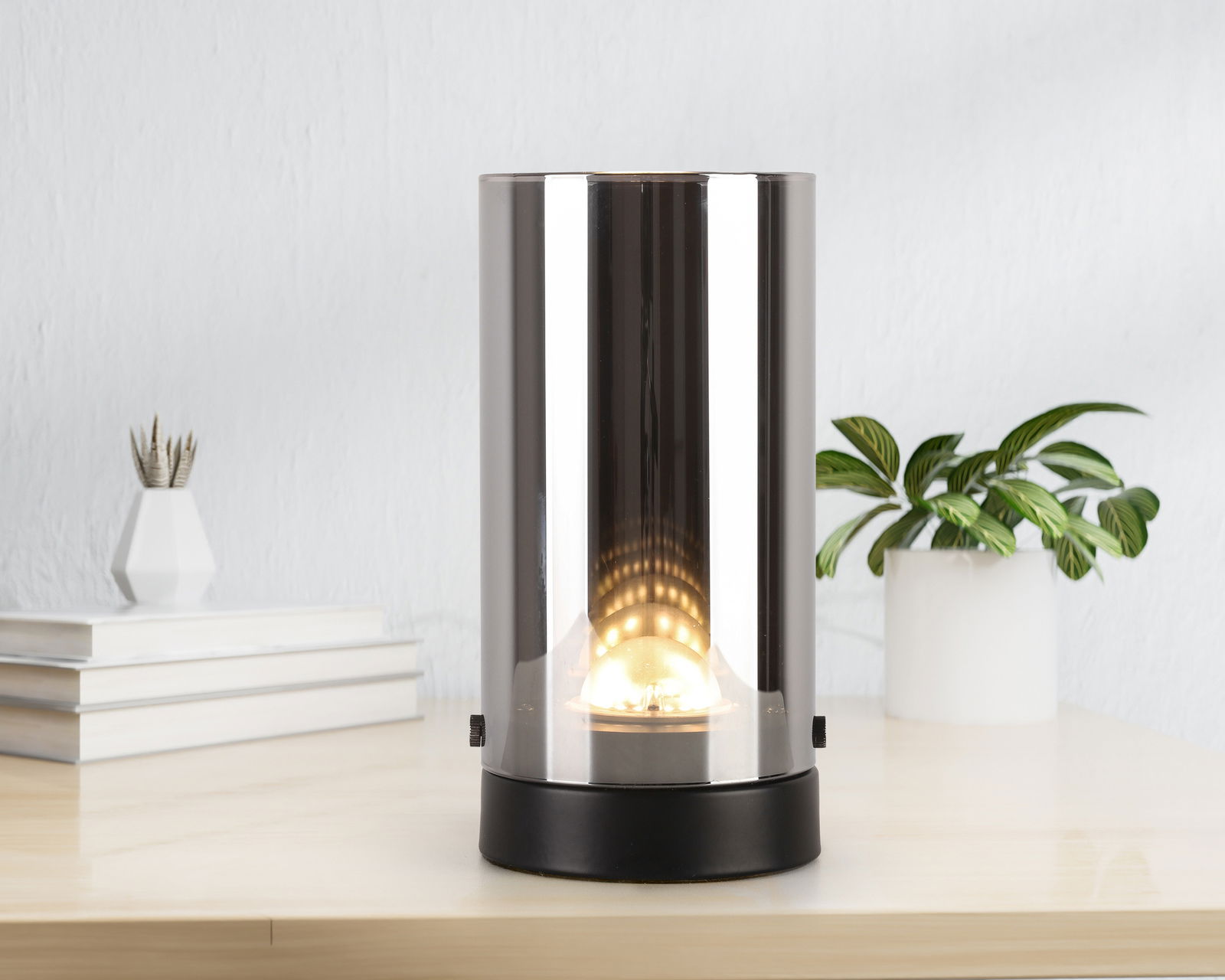 Zylindrische LED-Tischleuchte mit spiegelnder Oberfläche, frontal auf einem Tisch platziert, umgeben von Pflanzen und Büchern.