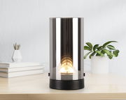 Zylindrische LED-Tischleuchte mit spiegelnder Oberfläche, frontal auf einem Tisch platziert, umgeben von Pflanzen und Büchern.