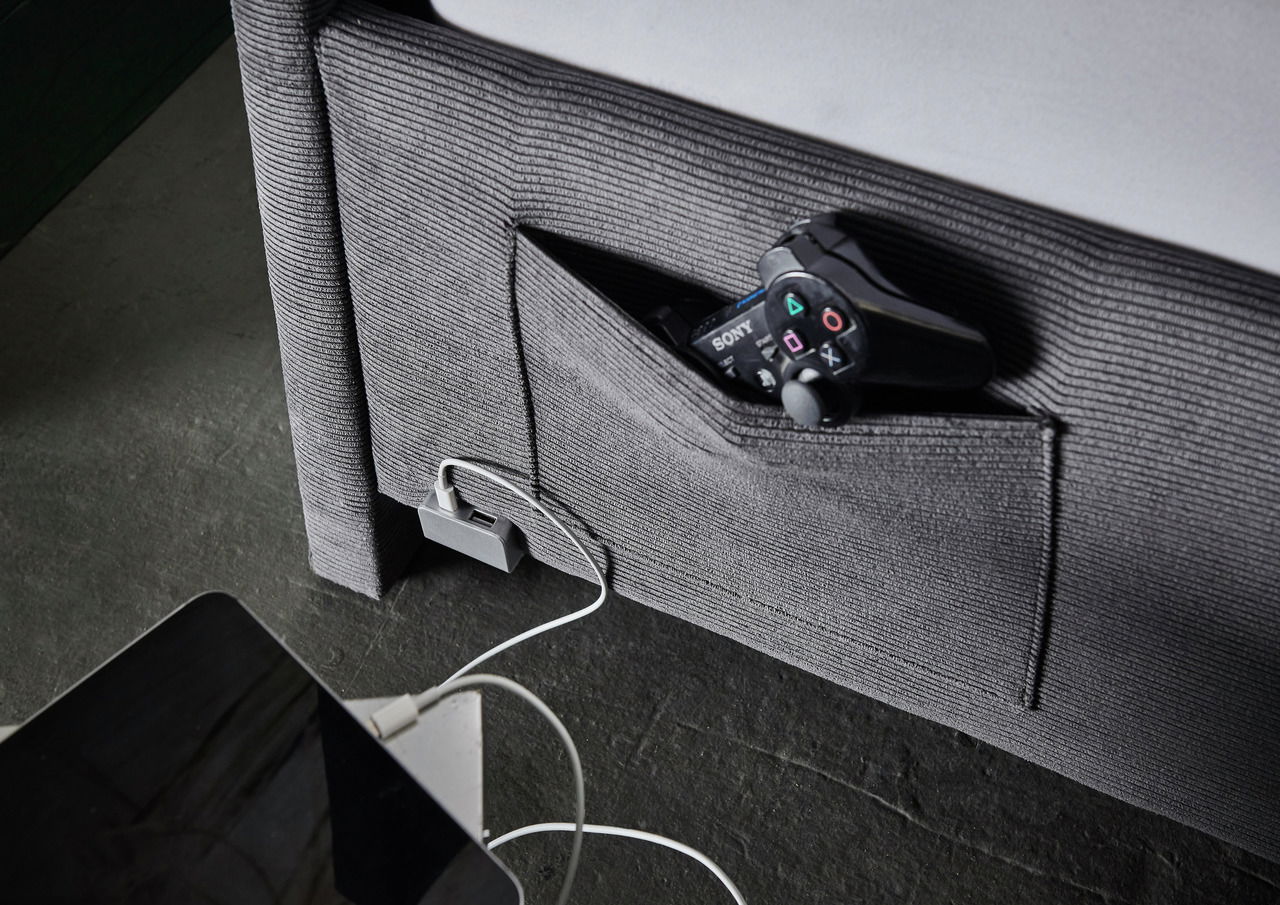 Nahaufnahme einer grauen Polsterliege mit integrierter Seitentasche, die einen Controller enthält, und einem USB-Ladeanschluss. Perspektive von der Seite.