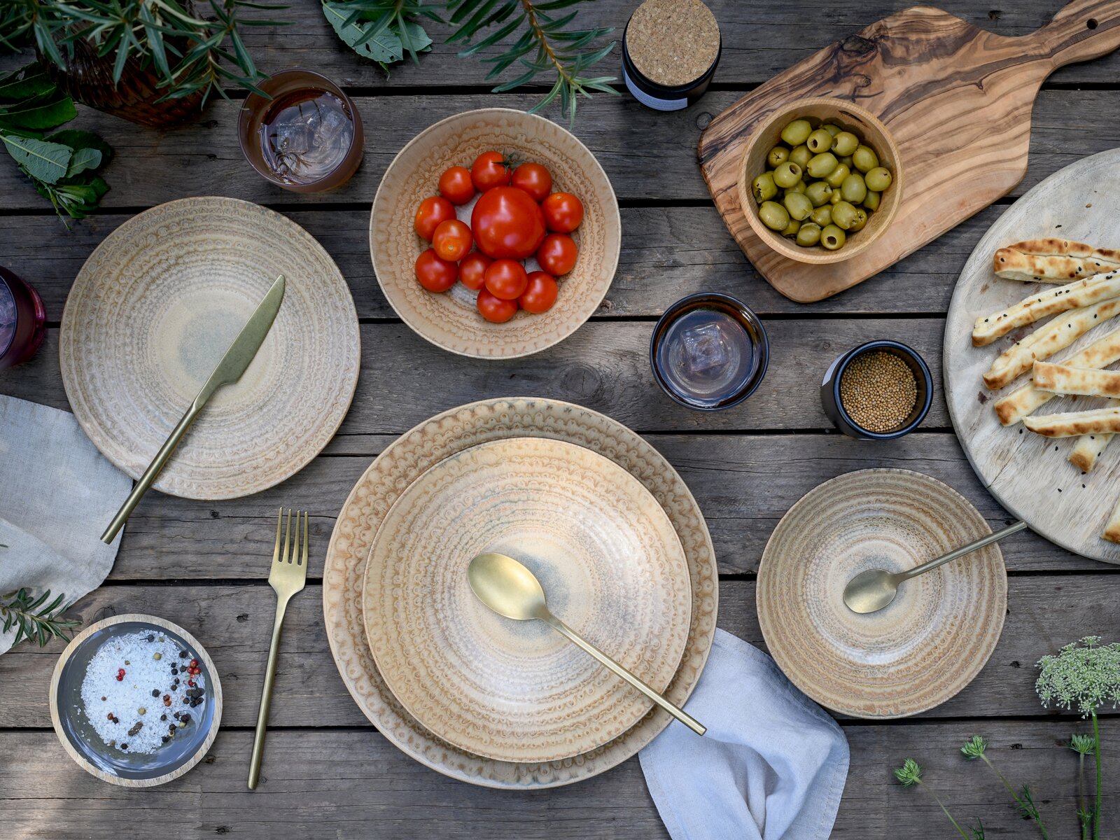 Creatable Tellerset 16-tlg NEAPEL Draufsicht auf rustikale Tischdeko: beigefarbenes Keramikgeschirr mit goldfarbenem Besteck auf Holztisch, dazu Schalen mit Tomaten und grünen Oliven, Brotstangen und Gewürze.