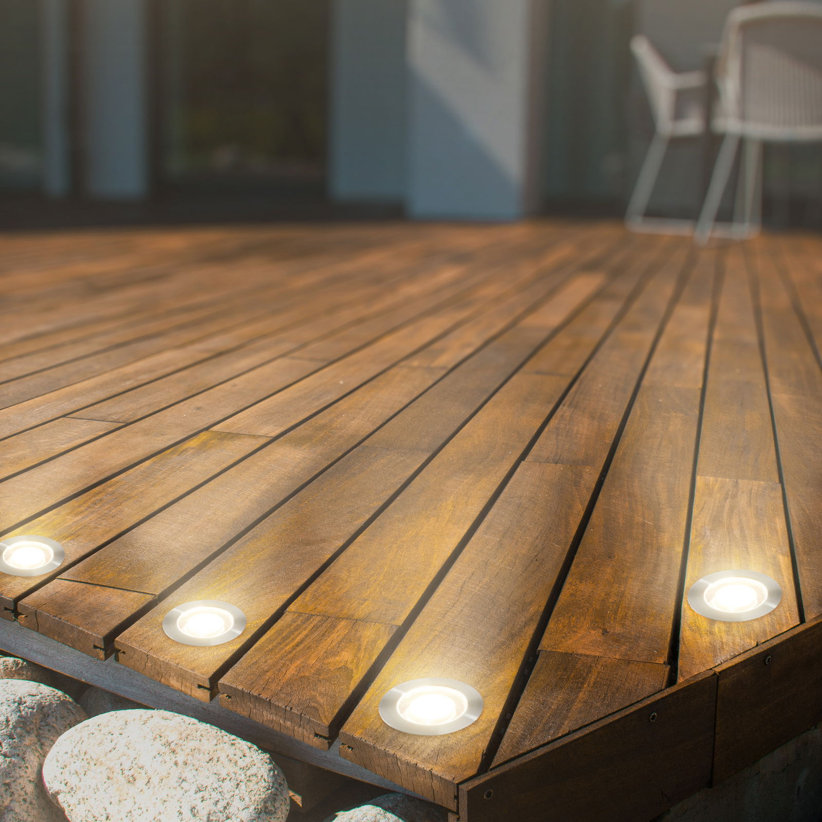 Perspektive von schräg oben auf eine Holzterrasse mit eingebauten Edelstahl-LED-Leuchten, die warmes Licht abstrahlen.