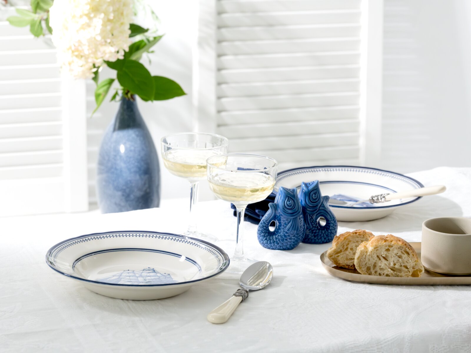 Maritimes Tischgedeck mit weiß-blauen Tellern, zwei Sektschalen, blauen Fischfiguren, Brot und Vase mit Blumen auf weißer Tischdecke, aus leicht erhöhter schräger Perspektive