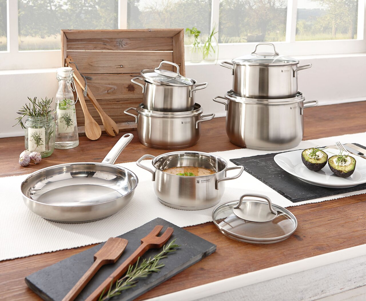 Topfset Brillant 6-tlg. aus Edelstahl, bestehend aus mehreren Töpfen und Pfannen mit Glasdeckeln, auf einem Holztisch in einer hellen Küche präsentiert. Perspektive: seitlich von vorne.