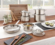 Topfset Brillant 6-tlg. aus Edelstahl, bestehend aus mehreren Töpfen und Pfannen mit Glasdeckeln, auf einem Holztisch in einer hellen Küche präsentiert. Perspektive: seitlich von vorne.