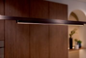 Moderne, schlanke LED-Pendelleuchte in Schwarz mit warmweißem Licht, seitliche Nahaufnahme in leicht schräger Perspektive vor unscharfem Wohnraum-Hintergrund