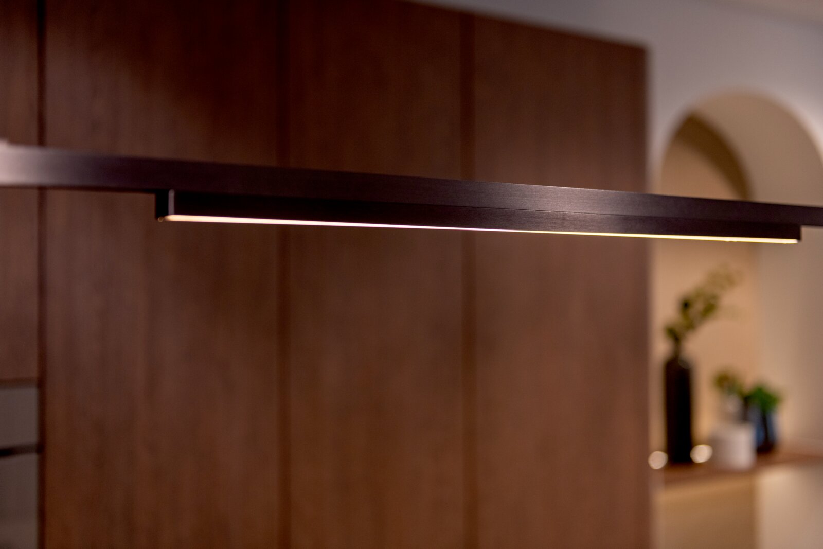 Moderne, schlanke LED-Pendelleuchte in Schwarz mit warmweißem Licht, seitliche Nahaufnahme in leicht schräger Perspektive vor unscharfem Wohnraum-Hintergrund