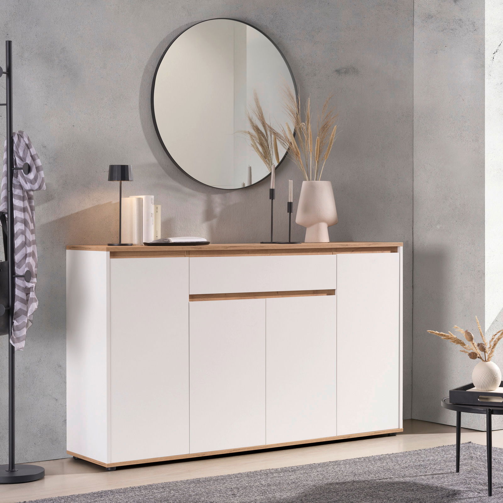 Modernes Sideboard in Weiß mit Holzdetails, dekoriert mit einer Lampe, Büchern und einer Vase, im Vordergrund ein runder Spiegel an der Wand, seitliche Perspektive.