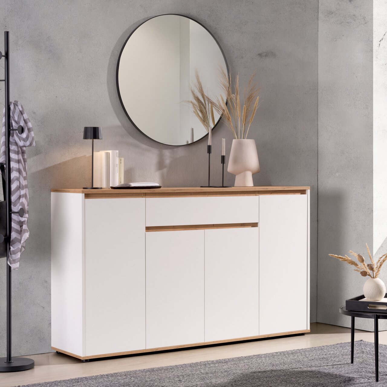 Carryhome XL-Sideboard MALEA Modernes Sideboard in Weiß mit Holzdetails, dekoriert mit einer Lampe, Büchern und einer Vase, im Vordergrund ein runder Spiegel an der Wand, seitliche Perspektive.