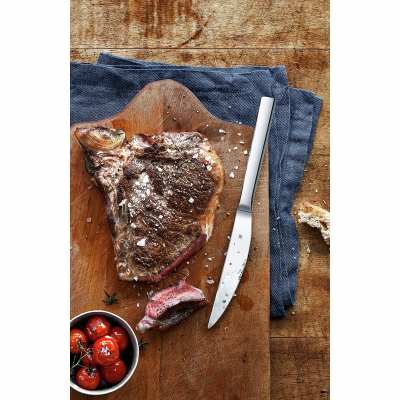 WMF Steakmesser 6er-Set NUOVA Steakmesser auf einem Holzbrett mit einem saftigen Steak und Kirschtomaten, von oben betrachtet.
