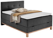 Graues Boxspringbett mit Holzfüßen, dekoriert mit Kissen und Decken, seitliche Perspektive