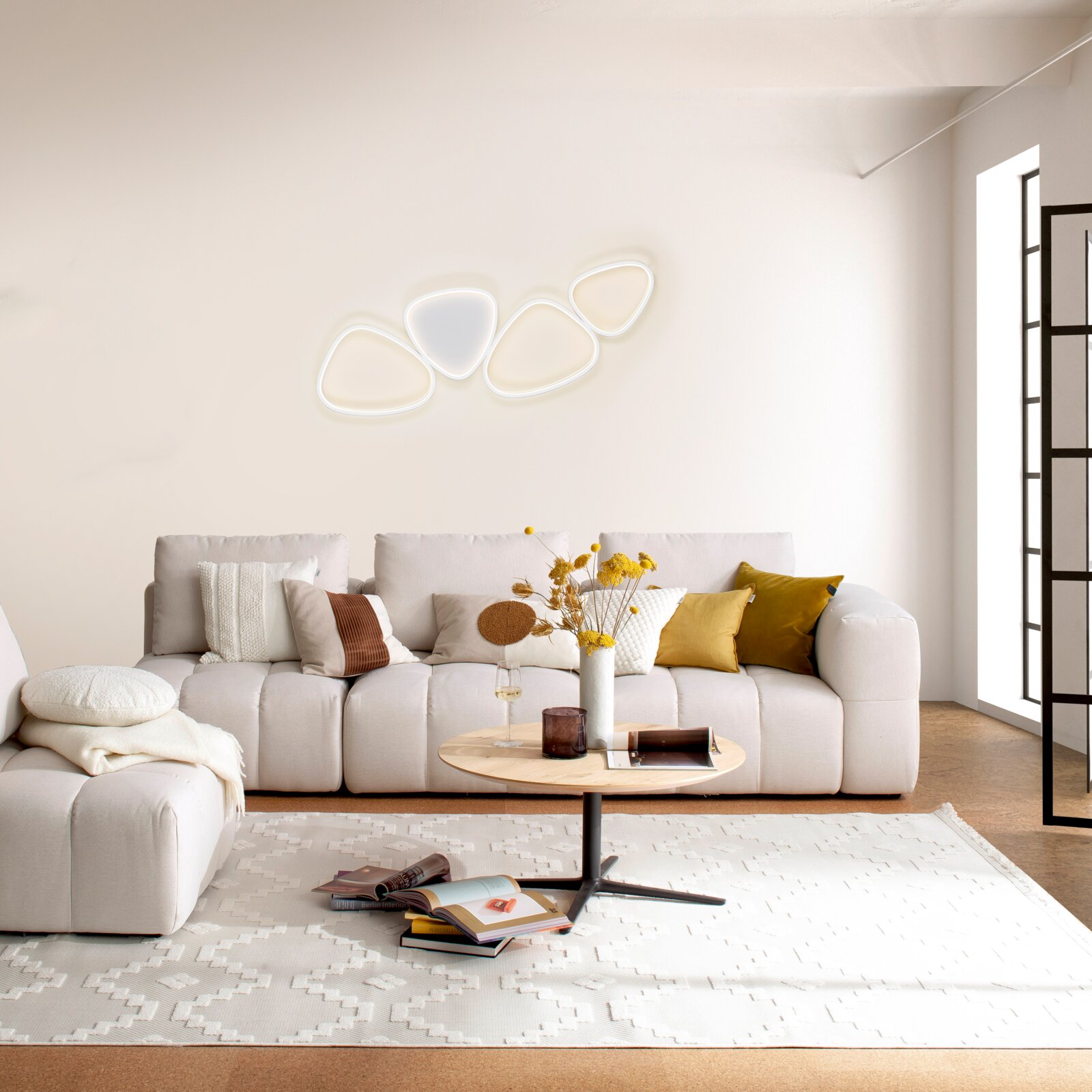 Modernes Wohnzimmer mit beigem Sofa und Couchtisch; an der Wand leuchtet eine abstrakte, mehrteilige LED-Wandleuchte in warmweiß, frontal aus leicht erhöhter Perspektive fotografiert.