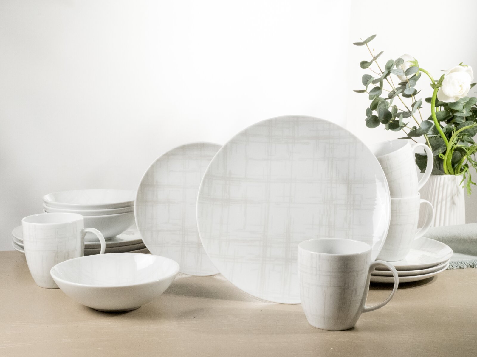 Weißes Geschirrset mit dezentem grauen Linienmuster (Teller, Schalen und Tassen) auf hellem Holztisch, frontal in leichter Seitenperspektive fotografiert im Hintergrund eine Vase mit Blumen und Grün.