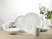 Weißes Geschirrset mit dezentem grauen Linienmuster (Teller, Schalen und Tassen) auf hellem Holztisch, frontal in leichter Seitenperspektive fotografiert; im Hintergrund eine Vase mit Blumen und Grün.