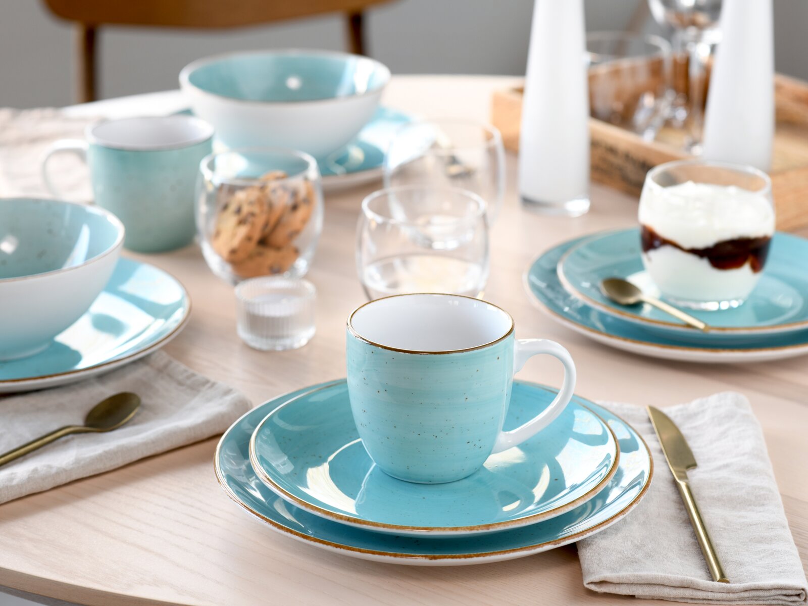 Hellblaues Keramik-Gedeck mit Tasse auf Untertasse und Teller, goldfarbenem Besteck und Frühstücksaccessoires auf Holztisch, Perspektive: leicht von oben in schräger Frontansicht