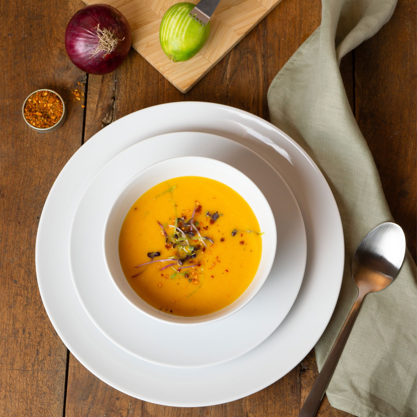 Vogelperspektive auf ein weißes Geschirrset mit einer Schüssel Suppe, umgeben von einer Zwiebel, einem geschnittenen Apfel, Gewürzen und einem Löffel auf einem Holztisch.