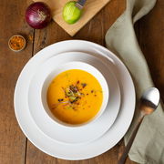 Vogelperspektive auf ein weißes Geschirrset mit einer Schüssel Suppe, umgeben von einer Zwiebel, einem geschnittenen Apfel, Gewürzen und einem Löffel auf einem Holztisch.