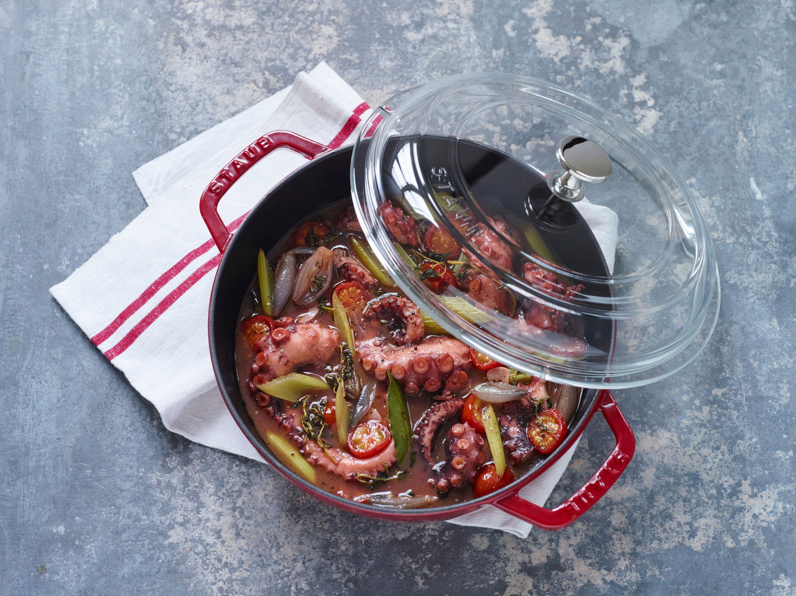 Roter Bräter mit Glasdeckel, gefüllt mit einem Eintopf aus Meeresfrüchten und Gemüse, von oben fotografiert.