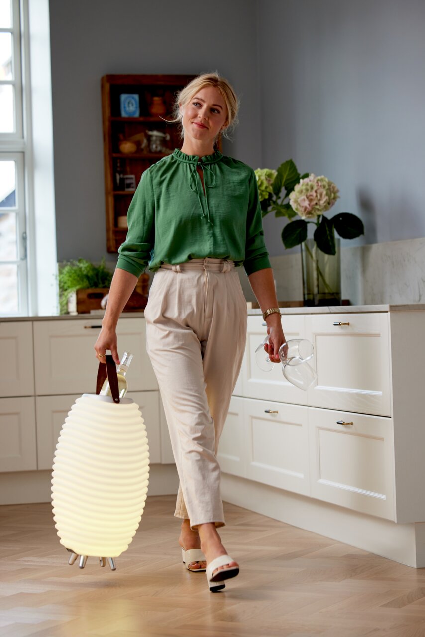 kooduu LED-Stehleuchte SYNERGY Frau in einer Küche trägt eine leuchtende Akku-Leuchte mit Lautsprecher in der Hand, seitliche Perspektive.