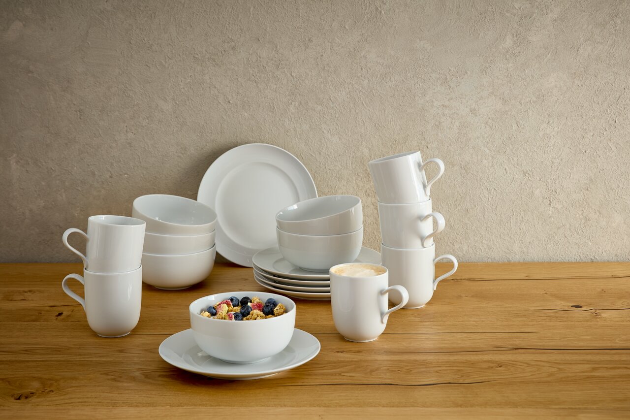Seltmann Weiden Frühstücksset 18-tlg. LIBERTY DELUXE Weißes, minimalistisches Porzellan-Geschirrset mit Tellern, Schalen und Bechern auf einem Holztisch vor heller Wand, seitliche Perspektive auf das Arrangement.
