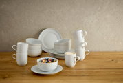 Weißes, minimalistisches Porzellan-Geschirrset mit Tellern, Schalen und Bechern auf einem Holztisch vor heller Wand, seitliche Perspektive auf das Arrangement.