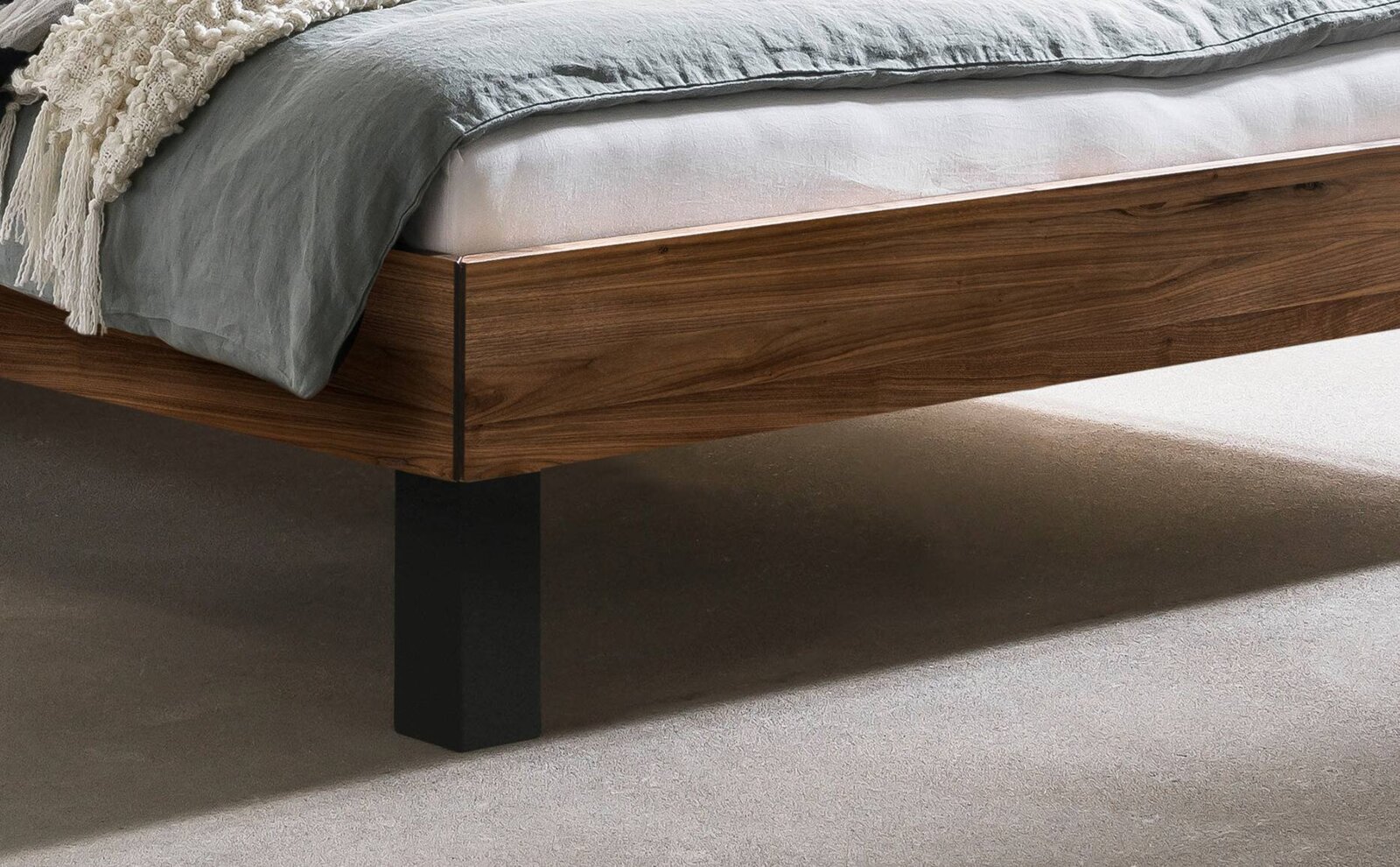 Detailfoto van een modern houten bed met een warm generfd bedrandje en een zwart metalen onderstel, met grijs beddengoed en een wit matras, gezien vanuit een zijdelingse hoek van onderaf