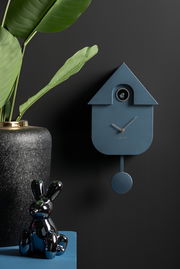Moderne blaue Wanduhr in Kuckucksuhrenform mit Pendel, seitliche Perspektive, dekoriert mit einer Pflanze und einer glänzenden Hasenfigur.
