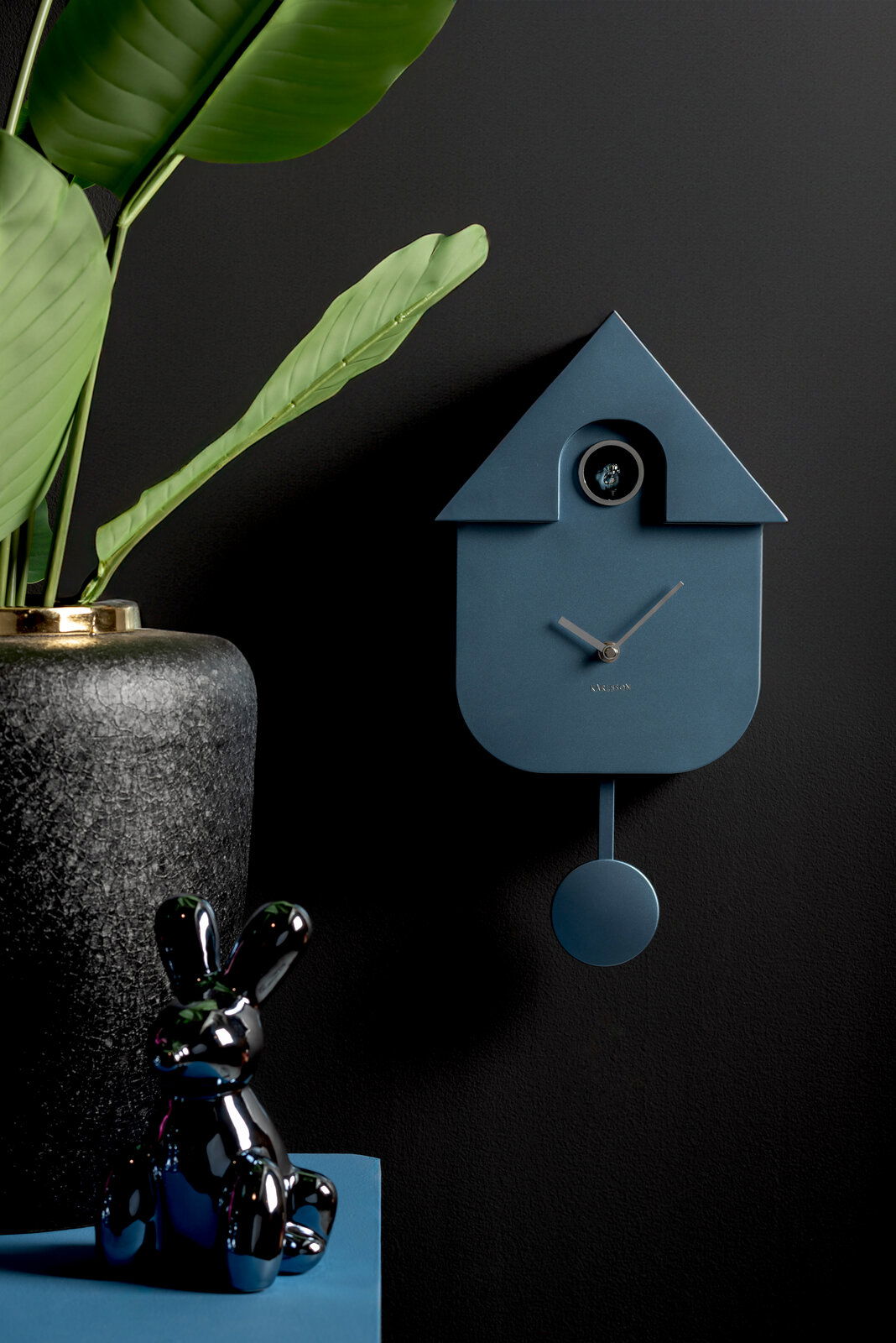 Moderne blaue Wanduhr in Kuckucksuhrenform mit Pendel, seitliche Perspektive, dekoriert mit einer Pflanze und einer glänzenden Hasenfigur.