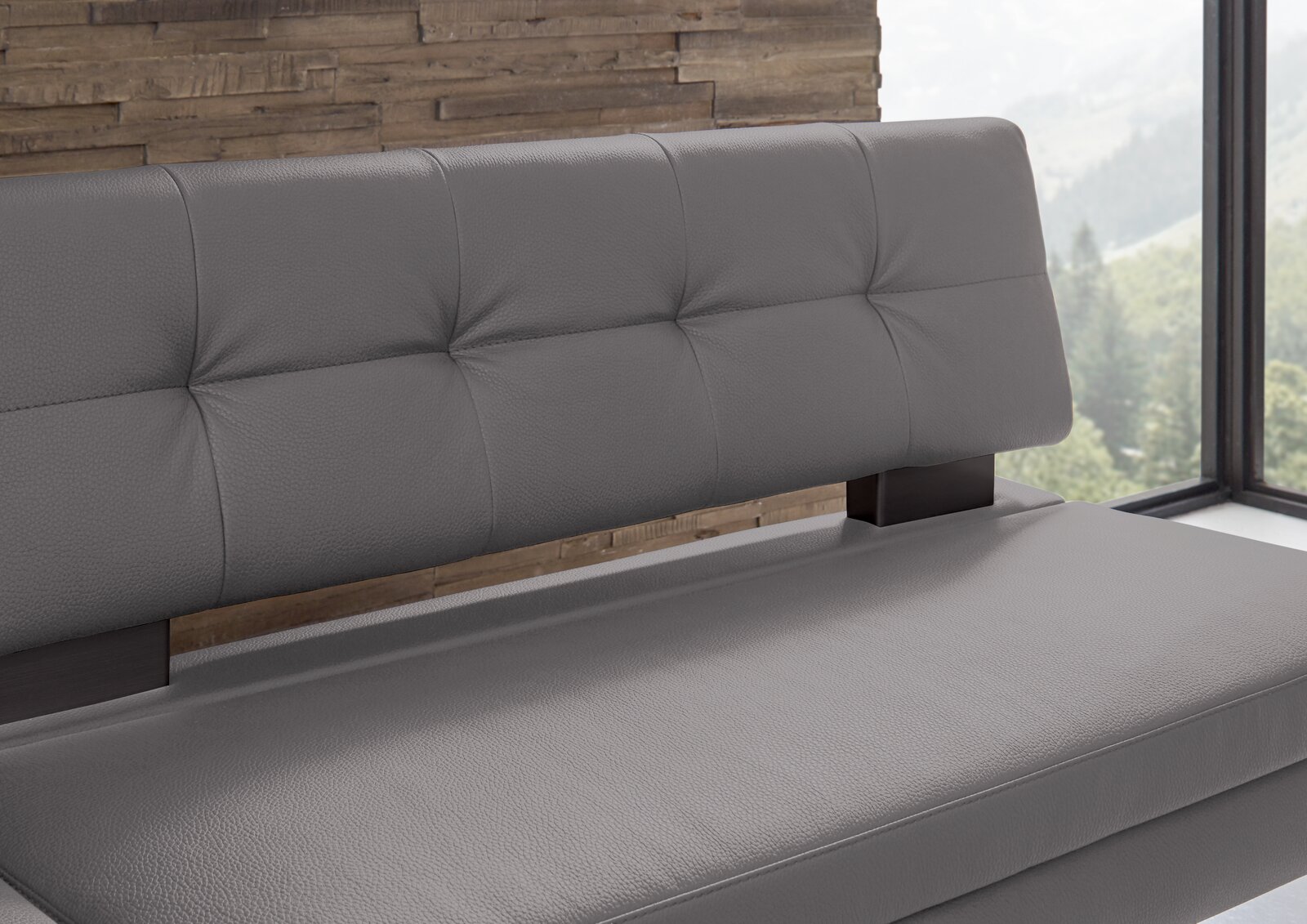 Graue Eckbank mit gepolsterter Rückenlehne und Sitzfläche, seitliche Perspektive, vor einer Holzwand und einem Fenster mit Blick ins Grüne.