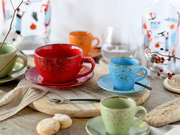 Bunte Keramik-Tassen mit Untertassen im gesprenkelten Design auf Holztisch, vorn eine große rote Tasse und eine kleine blaue Espressotasse; Perspektive: leicht von oben, frontnaher Blick