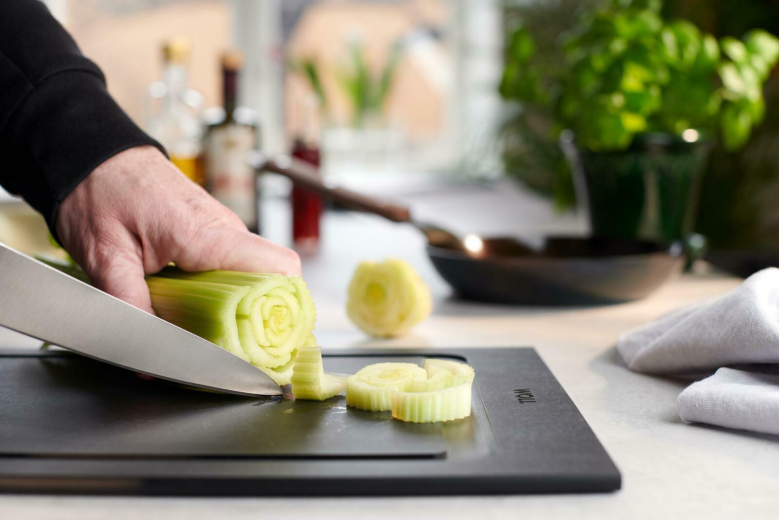 Nahaufnahme eines Schneidebretts mit einer Hand, die Sellerie schneidet. Im Hintergrund sind Küchenutensilien und Pflanzen zu sehen.