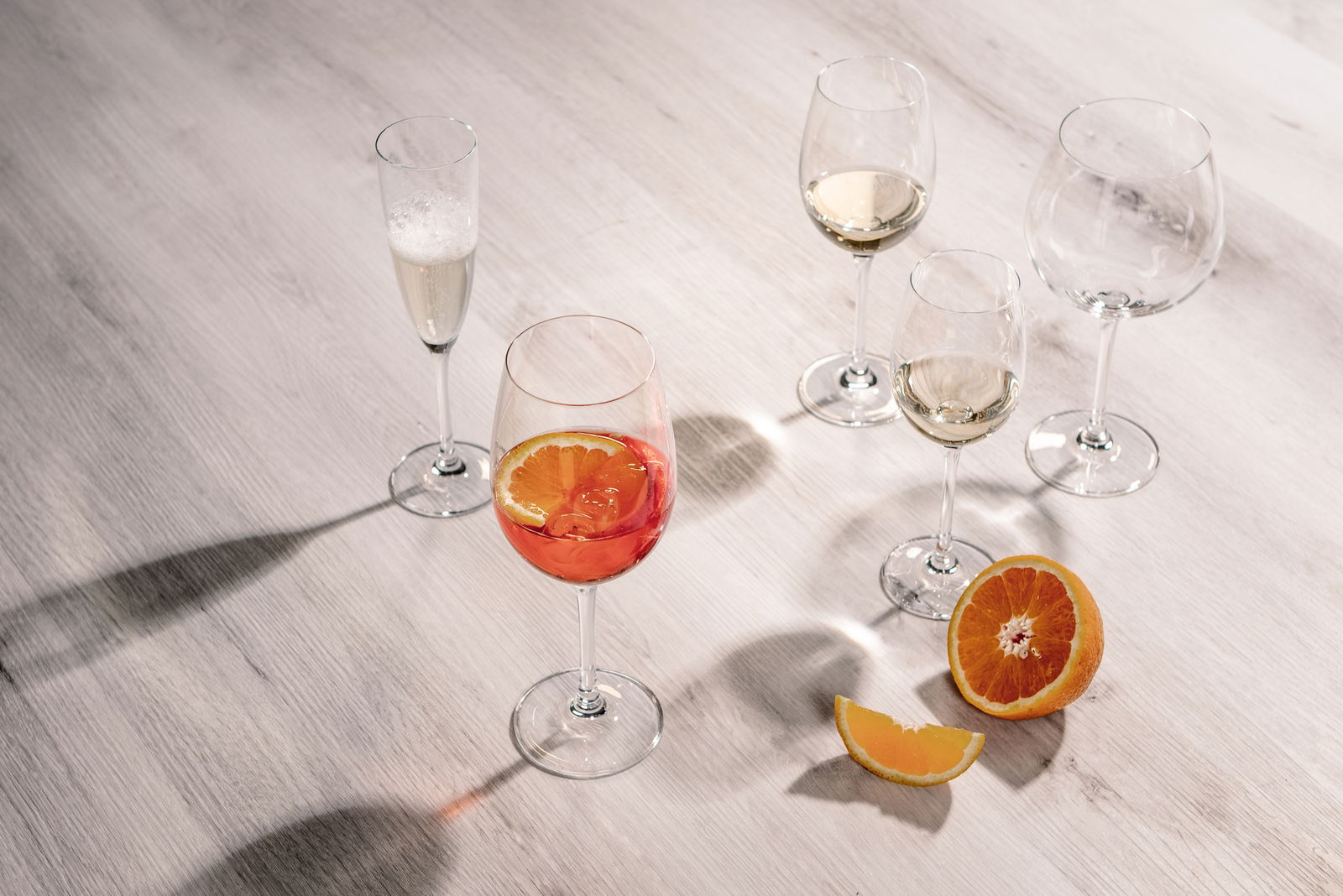 Fünf Weingläser auf einem Tisch, eines mit rotem Getränk und Orangenscheibe, aus der Vogelperspektive fotografiert.