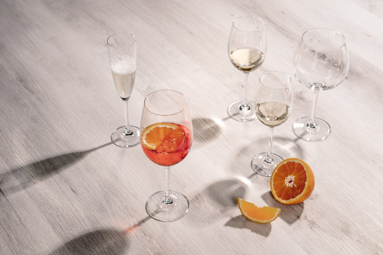 Fünf Weingläser auf einem Tisch, eines mit rotem Getränk und Orangenscheibe, aus der Vogelperspektive fotografiert.