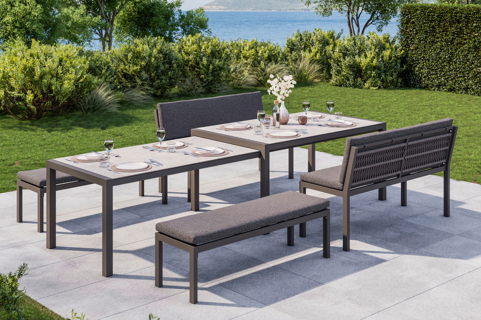 Lounge-Bank-Set auf einer Terrasse mit Blick auf einen Garten und das Meer, bestehend aus zwei Tischen, einer Bank und einer Sitzbank, gedeckt für ein Essen im Freien.