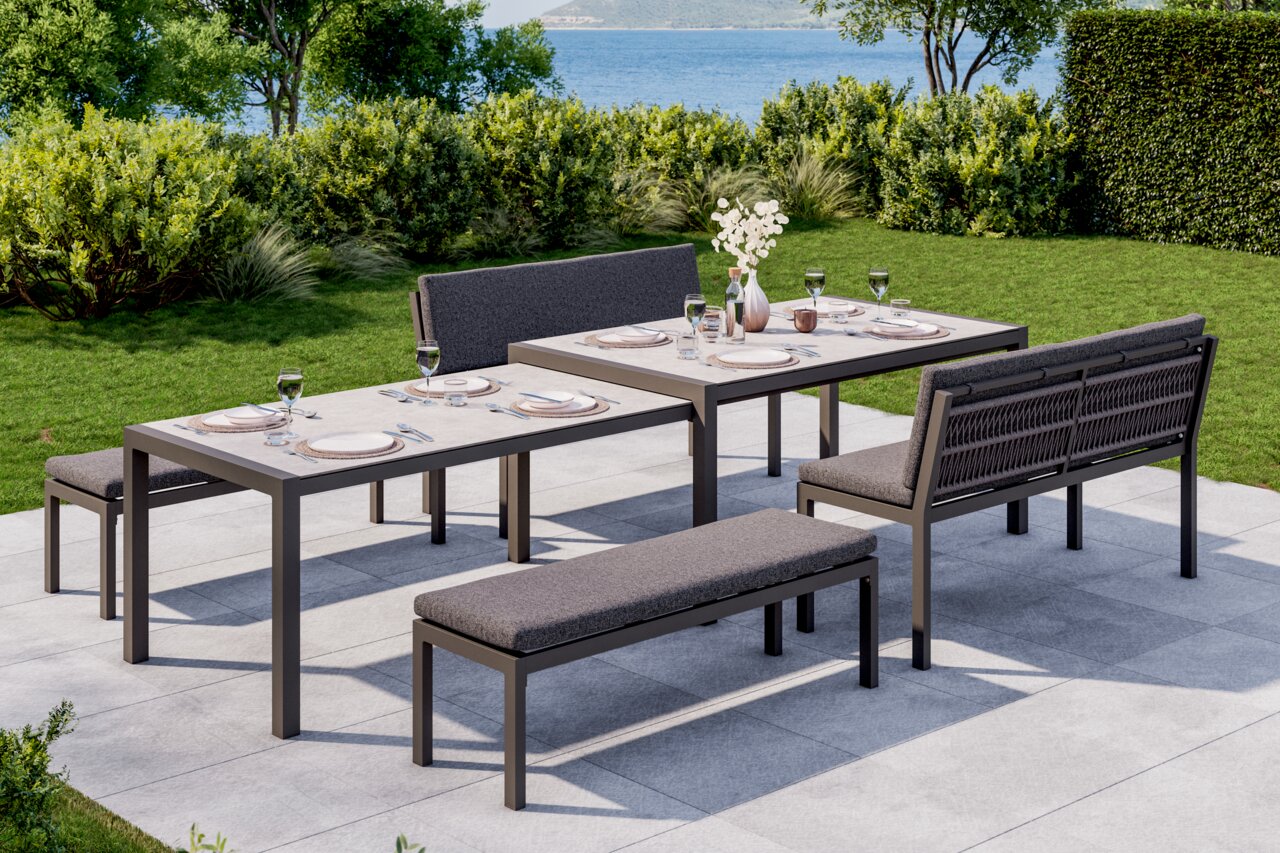 Lounge-Bank-Set auf einer Terrasse mit Blick auf einen Garten und das Meer, bestehend aus zwei Tischen, einer Bank und einer Sitzbank, gedeckt für ein Essen im Freien.