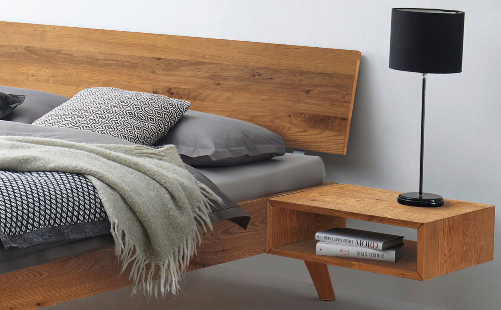 Holzbettgestell mit Kopfteil aus Holz, grauer Bettwäsche und Nachttisch mit schwarzer Lampe, seitliche Perspektive