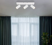 Deckenstrahler mit drei weißen, zylindrischen Leuchten, von unten betrachtet, vor einem Fenster mit Vorhängen.