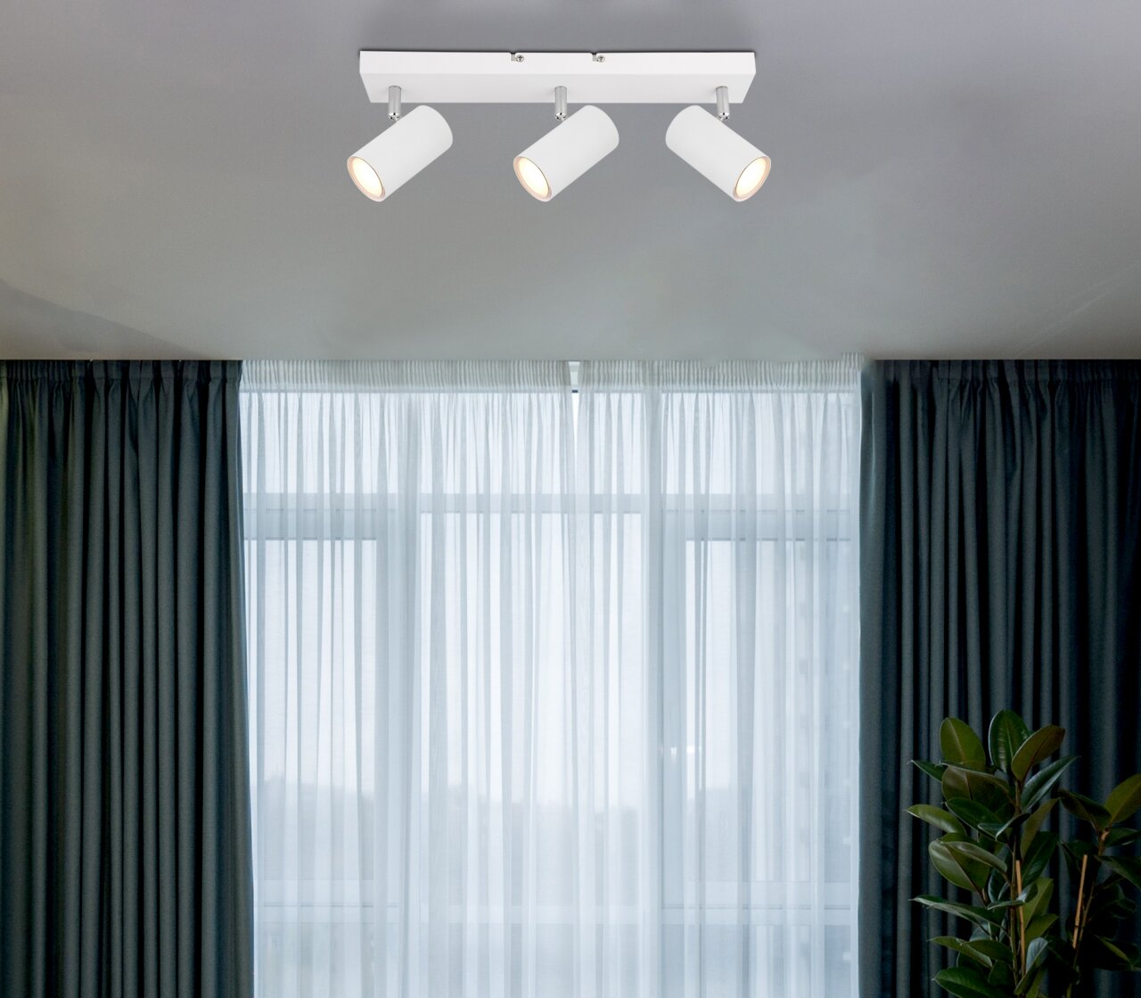 Deckenstrahler mit drei weißen, zylindrischen Leuchten, von unten betrachtet, vor einem Fenster mit Vorhängen.