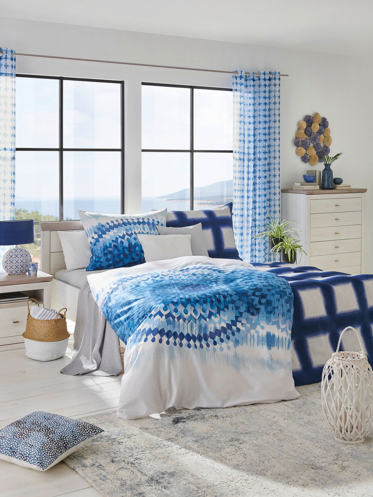 Modernes Schlafzimmer mit einem Bett 180x200, dekoriert mit blauer Bettwäsche und Kissen, in einem hellen Raum mit großen Fenstern und Blick auf das Meer, aus einer frontalen Perspektive.