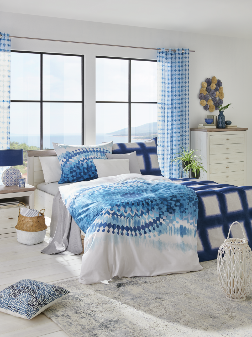 Modernes Schlafzimmer mit einem Bett 180x200, dekoriert mit blauer Bettwäsche und Kissen, in einem hellen Raum mit großen Fenstern und Blick auf das Meer, aus einer frontalen Perspektive.