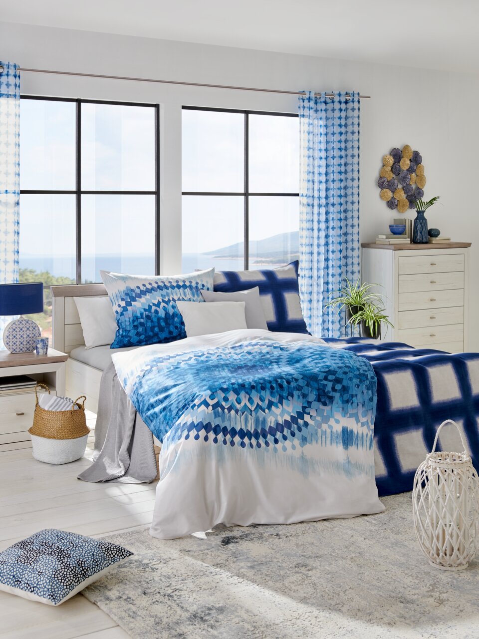 Modernes Schlafzimmer mit einem Bett 180x200, dekoriert mit blauer Bettwäsche und Kissen, in einem hellen Raum mit großen Fenstern und Blick auf das Meer, aus einer frontalen Perspektive.