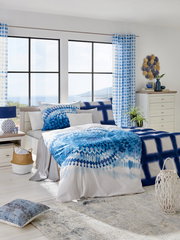 Modernes Schlafzimmer mit einem Bett 180x200, dekoriert mit blauer Bettwäsche und Kissen, in einem hellen Raum mit großen Fenstern und Blick auf das Meer, aus einer frontalen Perspektive.