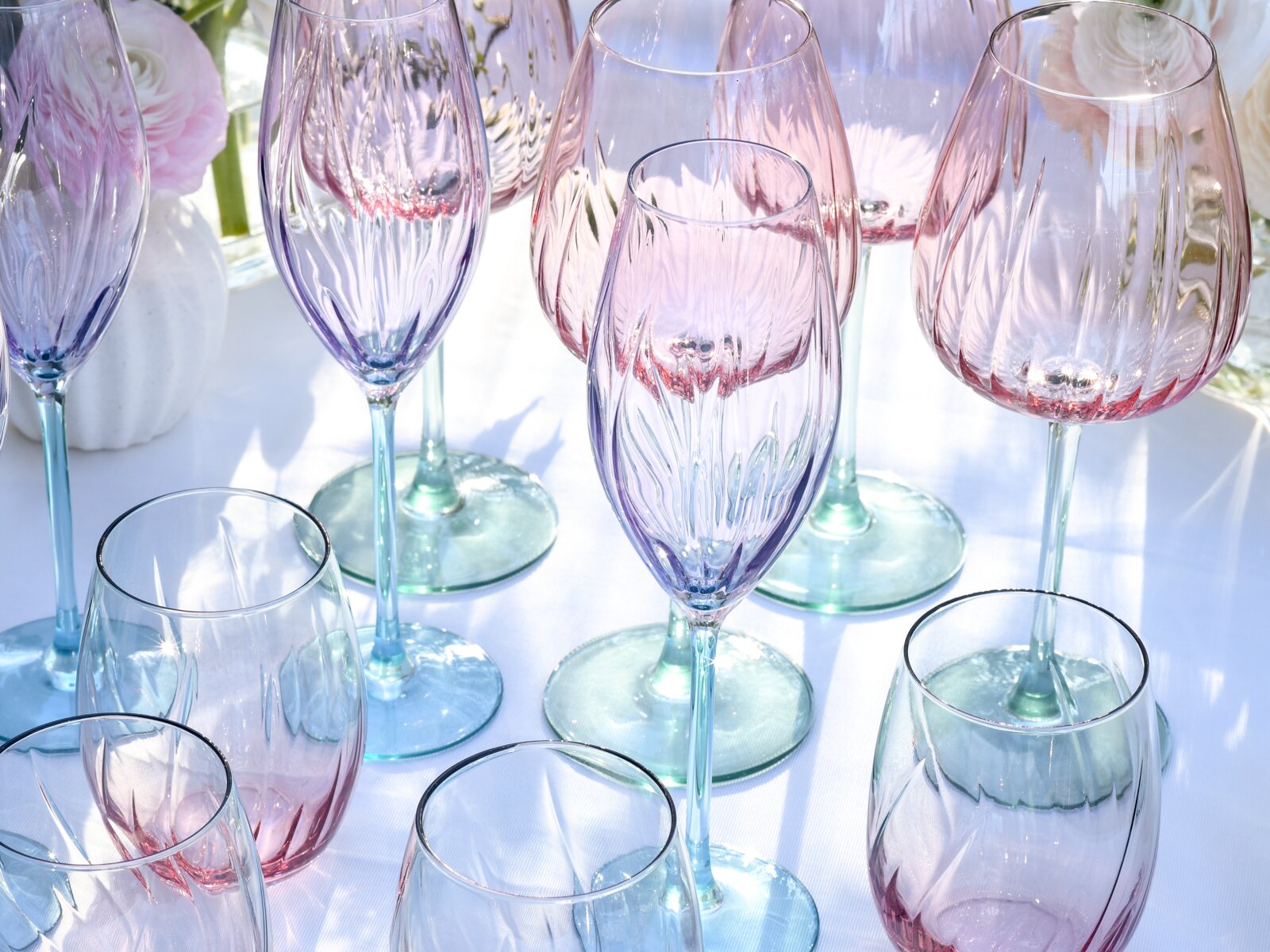 Nahaufnahme aus leicht erhöhter Perspektive: pastellfarbene Kristall-Gläser (Wein- und Sektgläser) in Rosa und Blau mit facettiertem Schliff auf weißer Tischdecke, im Sonnenlicht arrangiert.