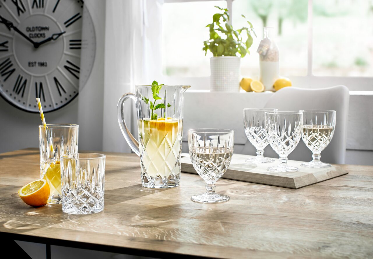 Een elegante Noblesse-set van 5 stuks op een tafel, met een kan en glazen van kristalglas, gevuld met drankjes, in een lichte ruimte met uitzicht op een raam.