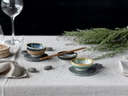 Schlicht gedeckter Tisch mit drei glasierten Keramikschälchen auf runden Schiefer-Untersetzern, Essstäbchen und Deko-Grün, fotografiert aus leicht erhöhter, schräger Seitenperspektive.