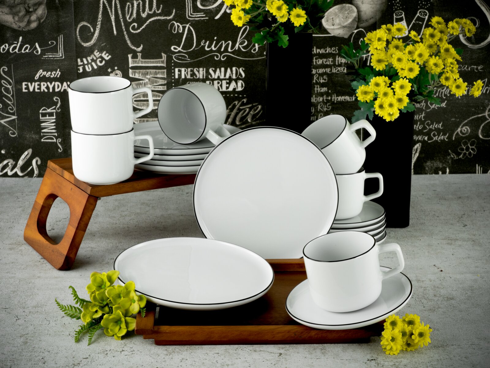 creaTable Kaffeeservice 18-tlg CHEF COLLECTION Weißes Porzellan-Geschirrset mit schwarzem Rand (Tassen, Untertassen, Teller und Schüssel) auf Holztabletts vor Tafelhintergrund mit gelben Blumen, fotografiert in leichter Aufsicht/3-4-Ansicht von vorn.
